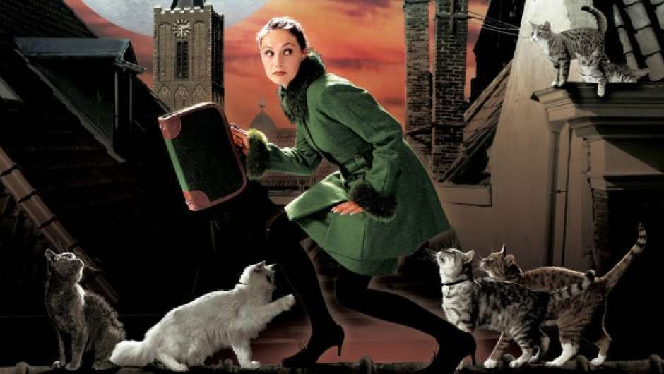 Eine Frau im grünen Mantel hält eine braune Aktentasche und geht geduckt, um sich guckend, übers Dach, umgeben von Katzen. 