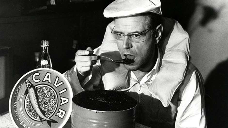 Ein Mann mit Schiebermütze und in einem weißen Daunenmantel, mit Brille, isst Kaviar aus der Dose. 