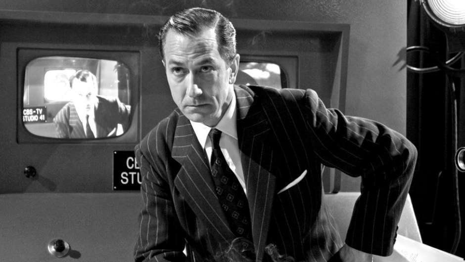 Schwarzweiß Bild, ein Mann im Nadelstreifenanzug, auf einem Hocker mit einem Bein kniend, an der Kamera vorbeischauend, im Hintergrund Fernsehmonitore