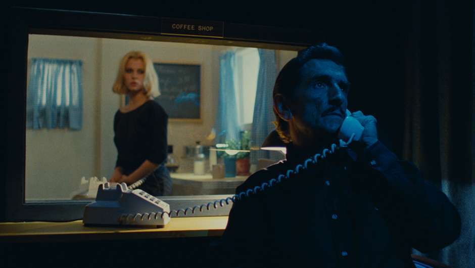Eine blonde Frau hinter einer Glasscheibe lauscht einem Mann, der in einen Telefonhörer spricht